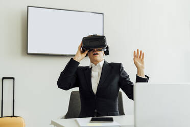 Mature female professional watching virtual reality simulator while sitting at desk in office - MEUF02285