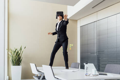 Frau benutzt ein Virtual-Reality-Headset, während sie auf einem Konferenztisch im Büro steht - MEUF02289