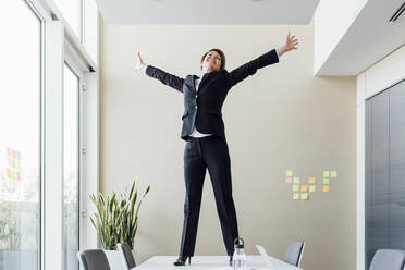 Happy businesswoman with arms raised celebrating success while standing on conference table in board room - MEUF02293