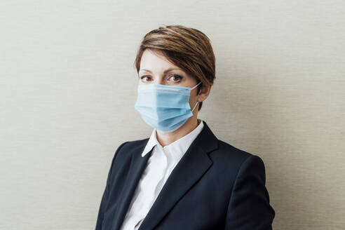 Female entrepreneur with protective face mask against beige wall in office - MEUF02294