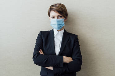 Businesswoman wearing protective face mask standing against beige wall in office - MEUF02296