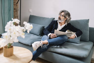 Hübscher älterer Mann mit Zeitung, der wegschaut, während er zu Hause auf dem Sofa sitzt - JCZF00544