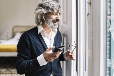 Lächelnder älterer Mann mit In-Ear-Kopfhörern, der in der Nähe des Fensters zu Hause mit seinem Smartphone telefoniert - JCZF00550