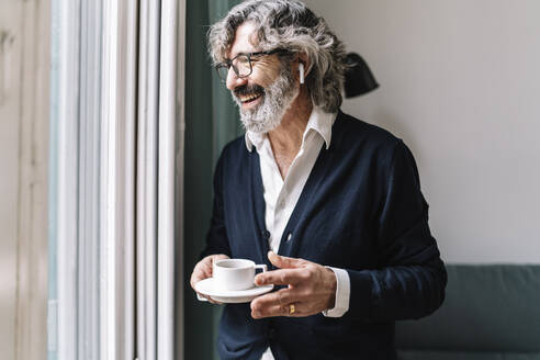 Glücklicher älterer Mann mit In-Ear-Kopfhörern, der eine Kaffeetasse in der Nähe des Fensters zu Hause hält - JCZF00551