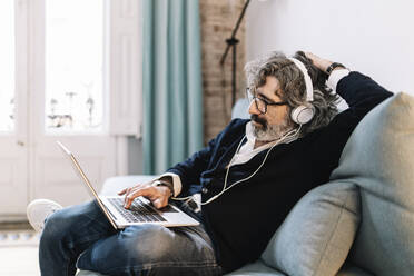 Entspannter älterer Mann mit Kopfhörern, der einen Laptop benutzt, während er zu Hause auf dem Sofa sitzt - JCZF00559