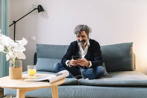 Lächelnder älterer Mann mit Kopfhörern, der ein Smartphone benutzt, während er zu Hause sitzt - JCZF00564