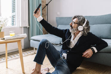 Mann mit Kopfhörern, der ein Selfie über sein Smartphone macht, während er zu Hause auf dem Boden sitzt - JCZF00567