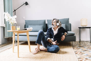 Nachdenklicher Mann mit Kopfhörern, der sein Smartphone hält, während er zu Hause sitzt - JCZF00570