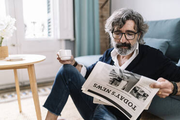 Hübscher Mann liest Zeitung und hält eine Kaffeetasse, während er zu Hause sitzt - JCZF00572