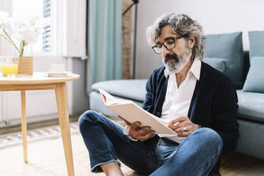 Älterer Mann liest ein Buch, während er zu Hause im Wohnzimmer sitzt - JCZF00574