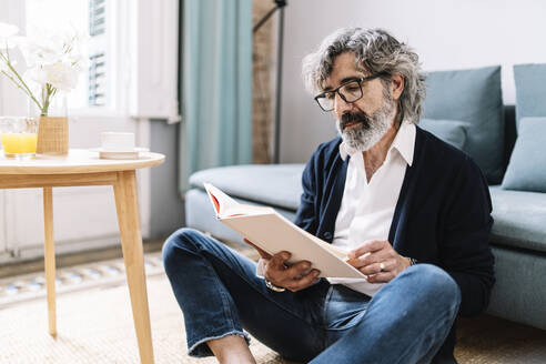 Älterer Mann liest ein Buch, während er zu Hause im Wohnzimmer sitzt - JCZF00574