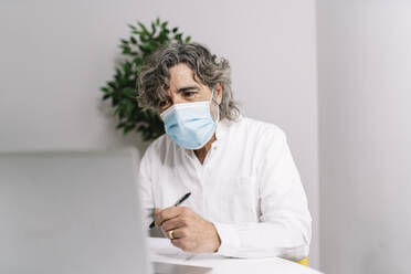 Businessman wearing protective face mask while working on laptop at office during COVID-19 - JCZF00577