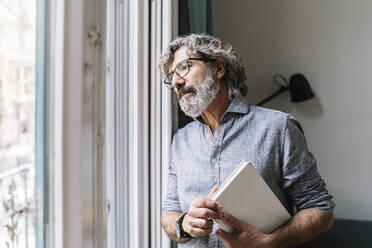 Nachdenklicher älterer Mann hält ein Buch in der Hand, während er durch das Fenster seines Hauses schaut - JCZF00582
