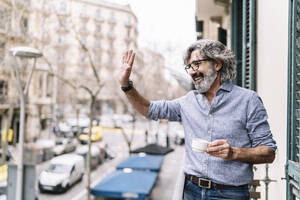 Fröhlicher älterer Mann winkt mit der Hand, während er auf dem Balkon eines Hauses Kaffee trinkt - JCZF00587