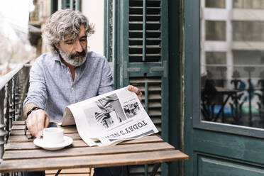 Mann trinkt Kaffee und liest Zeitung, während er am Tisch auf dem Balkon eines Hauses sitzt - JCZF00588