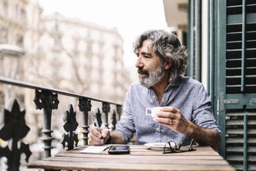 Älterer Mann, der Kaffee trinkt und in einen Notizblock schreibt, während er auf dem Balkon eines Hauses wegschaut - JCZF00590