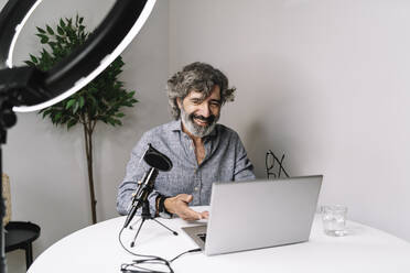 Senior male freelancer podcasting with laptop on table at home office - JCZF00593