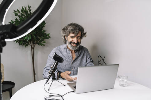 Älterer männlicher Freiberufler beim Podcasting mit Laptop auf dem Tisch im Heimbüro - JCZF00593