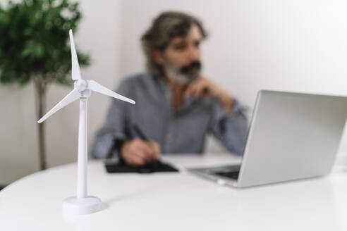 Wind turbine on table while businessman working in background at home office - JCZF00601