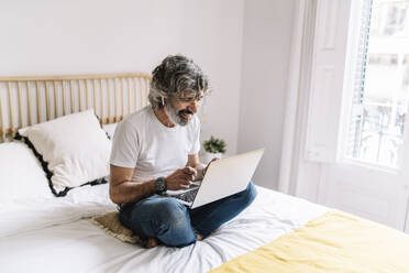 Lächelnder Mann, der einen Laptop benutzt, während er zu Hause auf dem Bett sitzt - JCZF00610