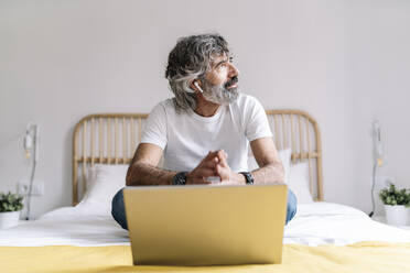 Thoughtful man looking away while sitting with laptop on bed at home - JCZF00613