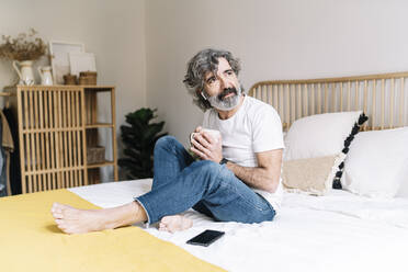 Nachdenklicher älterer Mann, der wegschaut und eine Kaffeetasse hält, während er zu Hause auf dem Bett sitzt - JCZF00615