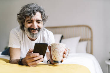 Happy senior man using smart phone holding coffee mug while lying on bed at home - JCZF00616