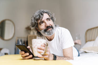 Mann mit Smartphone und Kaffeetasse schaut weg, während er zu Hause auf dem Bett liegt - JCZF00617