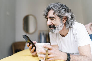 Man using mobile phone holding coffee mug while lying at home - JCZF00619