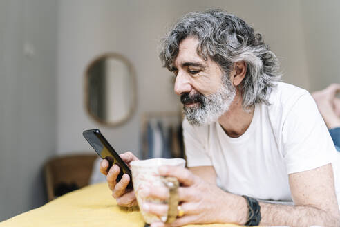 Mann, der ein Mobiltelefon benutzt und einen Kaffeebecher hält, während er zu Hause liegt - JCZF00619