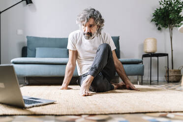 Man doing yoga on carpet while watching tutorial on laptop at home - JCZF00621