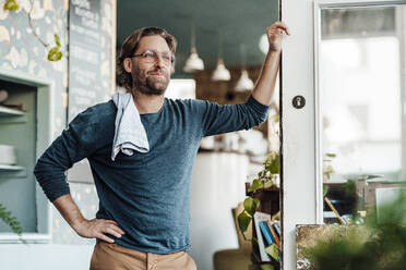 Älterer männlicher Besitzer mit Hand auf der Hüfte, der sich über die Tür eines Cafés lehnt - JOSEF03908