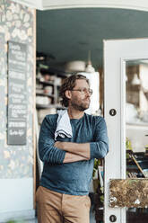 Männlicher Besitzer mit verschränkten Armen, der an der Tür eines Cafés lehnt - JOSEF03909