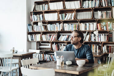 Männlicher Unternehmer mit Laptop vor einem Bücherregal im Café sitzend - JOSEF03924