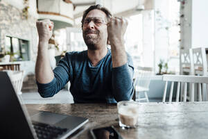 Happy male entrepreneur doing fist by laptop in coffee shop - JOSEF03949