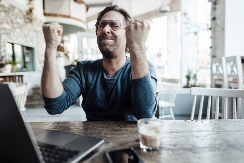 Happy male entrepreneur doing fist by laptop in coffee shop - JOSEF03949