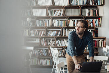 Man with laptop leaning on table in front of bookshelf at coffee shop - JOSEF03953