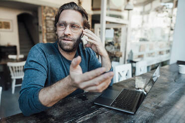 Männlicher Unternehmer, der in einem Café mit seinem Laptop telefoniert - JOSEF03960