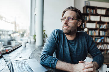 Thoughtful man looking away by laptop in coffee shop - JOSEF03962