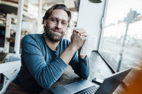 Lächelnder, selbstbewusster Mann am Laptop in einem Café während der COVID-19 - JOSEF03967