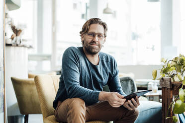 Smiling man with digital tablet sitting on sofa at cafe - JOSEF03992