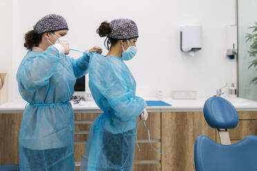 Female dentist helping while tying operating gown of colleague in clinic - EGAF02148