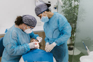 Orthodontists in protective workwear and face mask checking patient while standing by assistant in clinic - EGAF02149