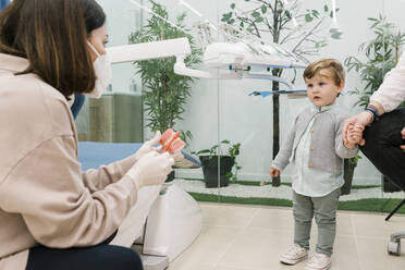 Kinderzahnarzt mit Gesichtsmaske, der einem Jungen mit seiner Mutter in der Klinik das Zähneputzen anhand eines Modells mit künstlichen Zähnen beibringt - EGAF02158