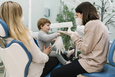 Kinderzahnarzt mit Gesichtsschutzmaske im Gespräch mit einem Jungen, der mit seiner Mutter in der Klinik sitzt - EGAF02162