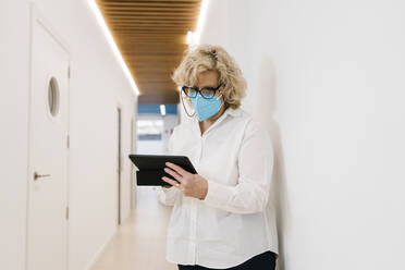 Mature dentist with protective face mask using digital tablet at clinic corridor - EGAF02167
