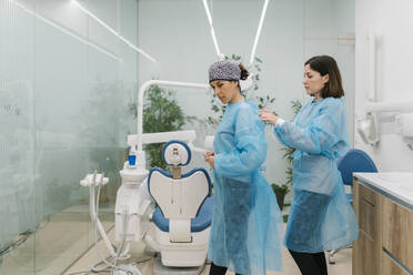 Female dentist helping assistant getting dressed in protective workwear at clinic - EGAF02185