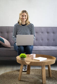 Blonde Mid Adult Frau sitzt mit Laptop auf dem Sofa - GIOF11982