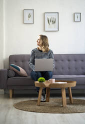 Thoughtful woman smiling while sitting with laptop on sofa - GIOF11984