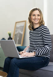 Lächelnde Frau im mittleren Erwachsenenalter, die zu Hause mit einem Laptop sitzt - GIOF11985